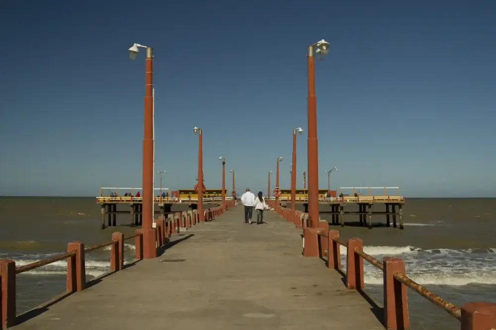 Pareja caminando por un muelle de cemento con faroles rojos que entra al mar, con pescadores en la plataforma del fondo