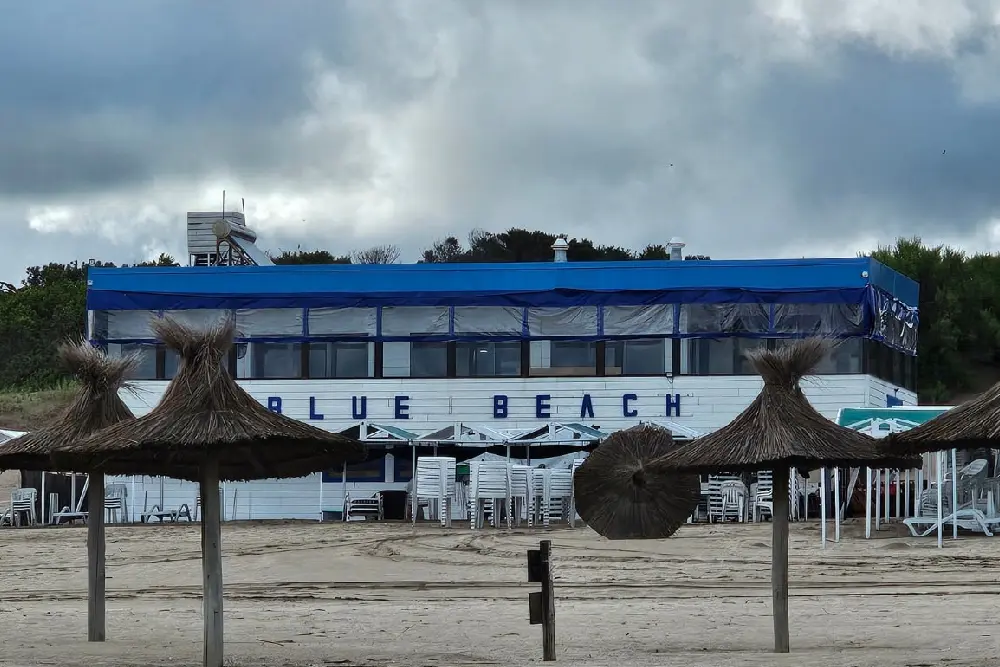 Edificio blanco con techo azul y el cartel Blue Beach sobre la arena, con sombrillas de paja en primer plano y el bosque detrás