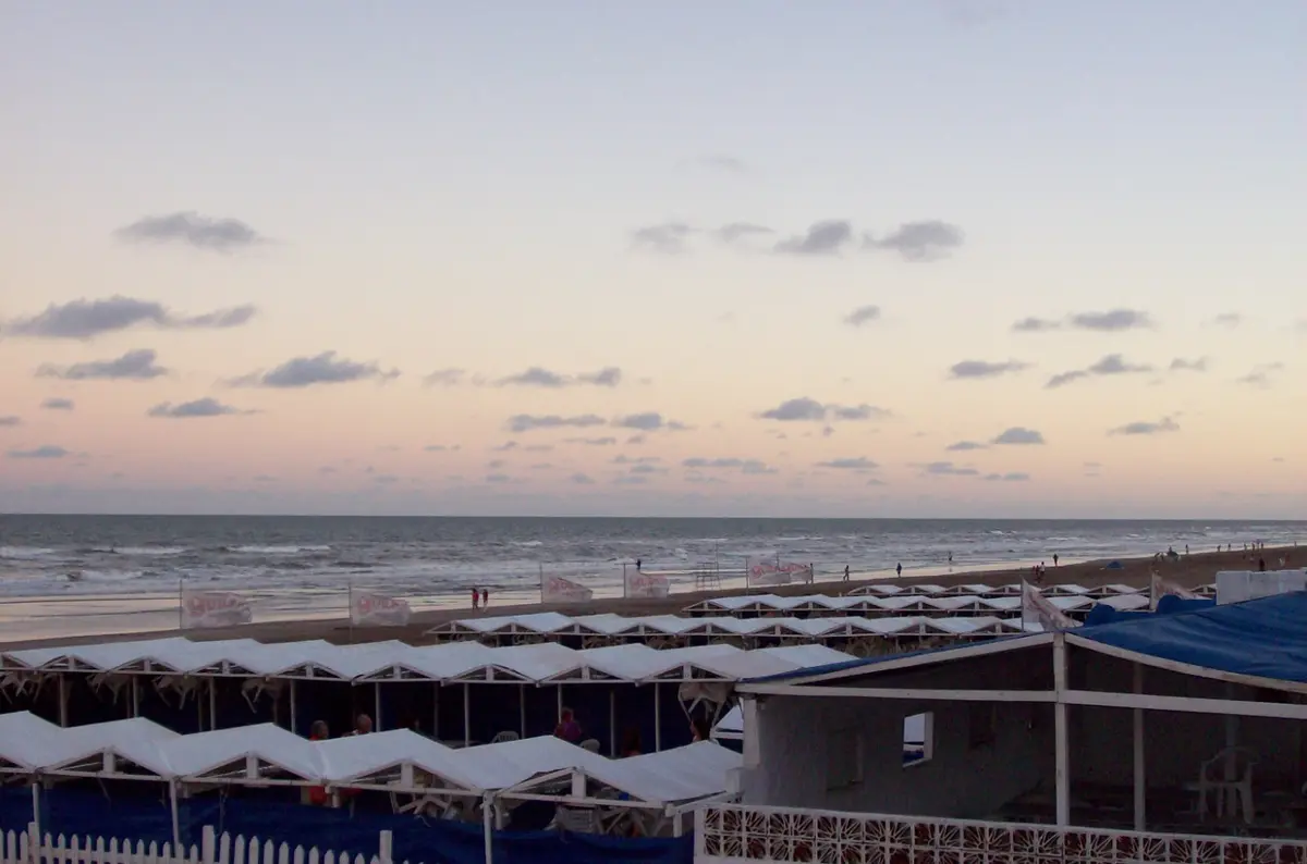 Hilera de carpas blancas de un balneario frente al mar al atardecer, con el cielo rosado y gente caminando por la orilla