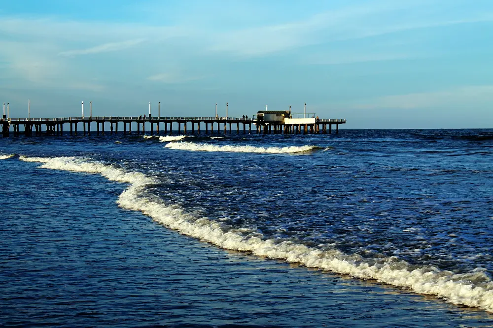 Muelle largo de madera sobre pilotes que entra al mar, con una ola rompiendo en primer plano y el cielo celeste