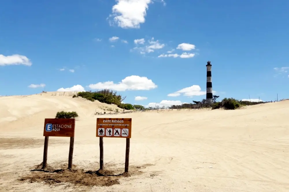 El Faro Querandí, de franjas blancas y negras, detrás de un médano, con carteles de estacionamiento y de prevención clavados en la arena