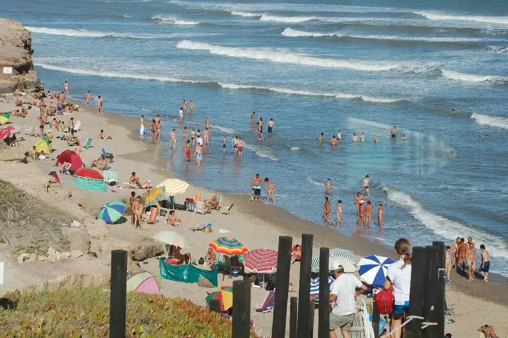 Playa angosta al pie de una barranca, con sombrillas de colores, mucha gente en la arena y bañándose, y olas al fondo