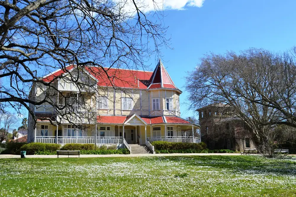 Casa de madera amarilla de dos plantas con techos rojos y galería, en medio de un parque con césped y árboles sin hojas