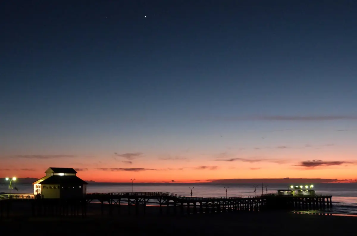 Silueta del muelle de madera con su casilla iluminada en la entrada, recortada contra el cielo naranja del atardecer