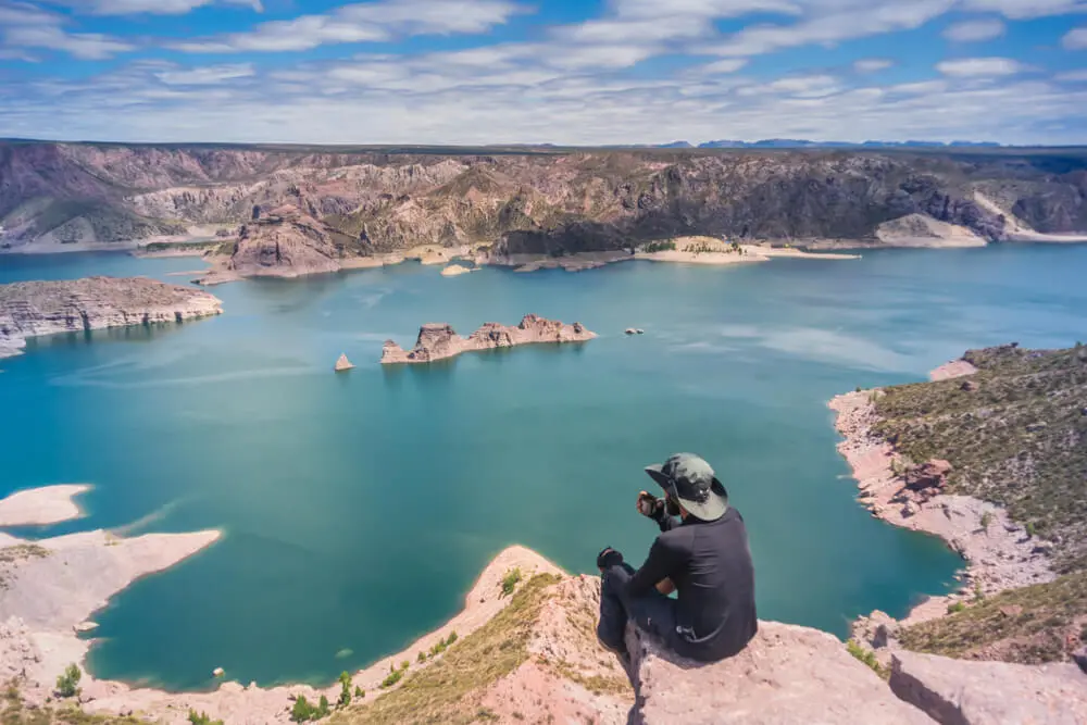 Un viajero sentado en una roca mira un embalse turquesa rodeado de barrancas de piedra rosada, con islotes en el agua
