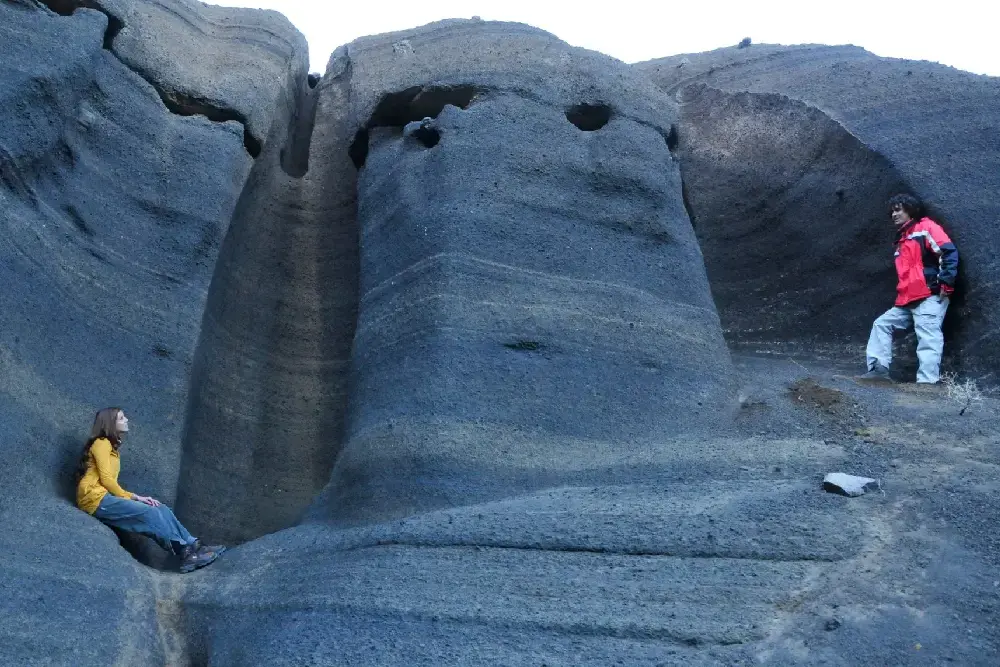 Dos personas junto a paredes de ceniza volcánica gris erosionadas en formas redondeadas, con huecos en la roca