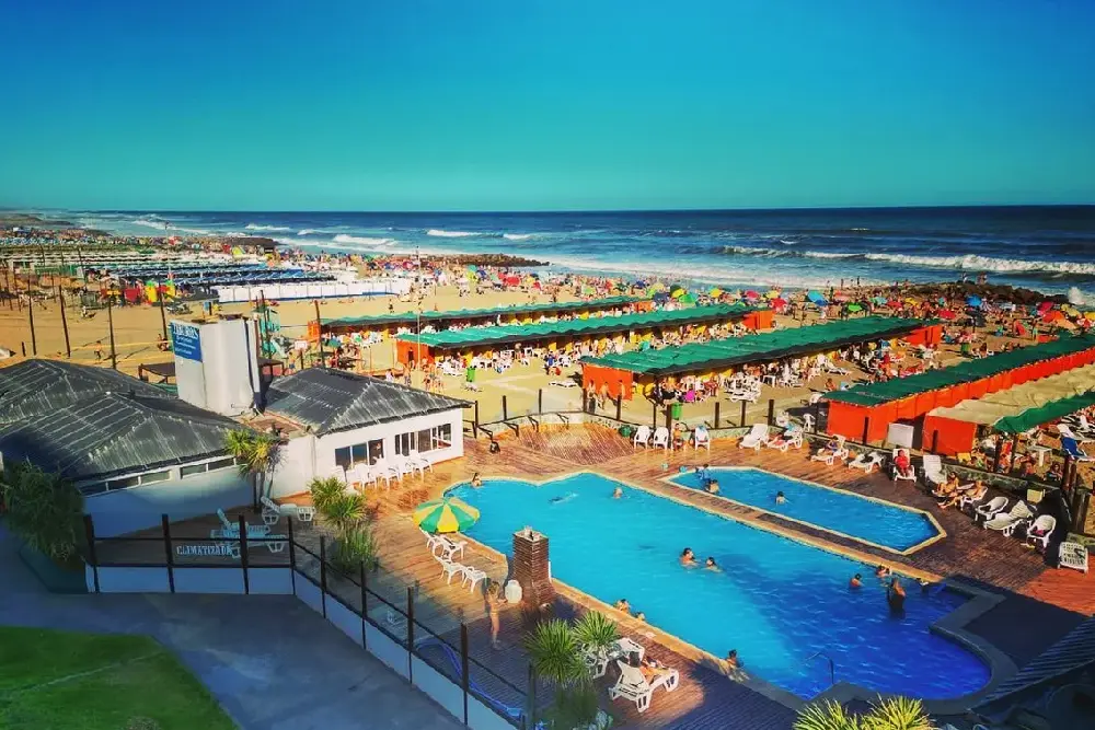 Balneario con dos piletas, reposeras blancas e hileras de carpas naranjas y verdes frente a una playa llena de sombrillas
