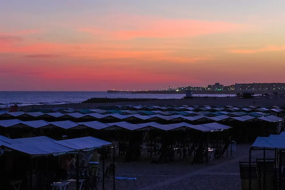 Atardecer rosado sobre la playa de Miramar, con hileras de carpas en primer plano y el muelle iluminado entrando al mar