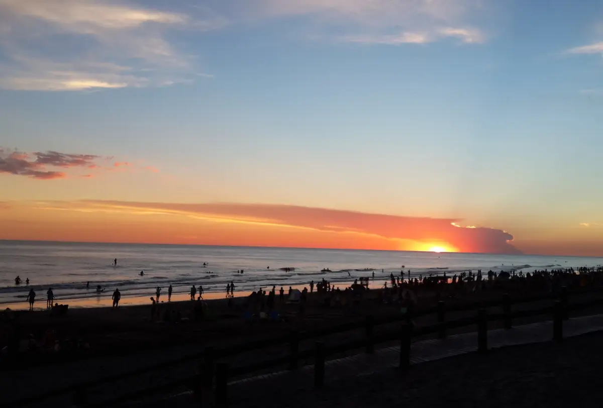 Atardecer sobre el mar con el sol tocando el horizonte, mucha gente en la orilla y la baranda de madera de la rambla en primer plano
