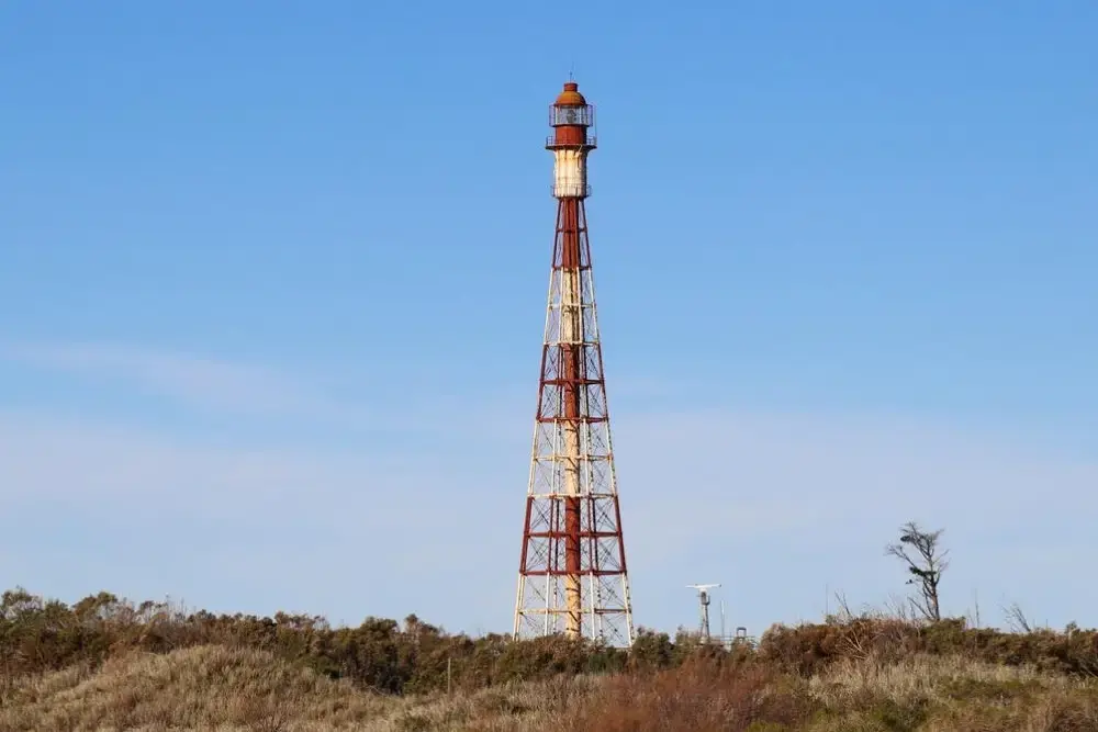 Torre metálica roja y blanca del Faro Recalada sobre médanos con vegetación baja y el cielo celeste