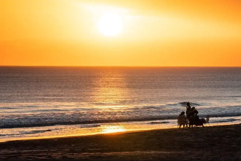 Un grupo sentado en reposeras en la orilla, frente al mar, con el sol bajo y el cielo anaranjado