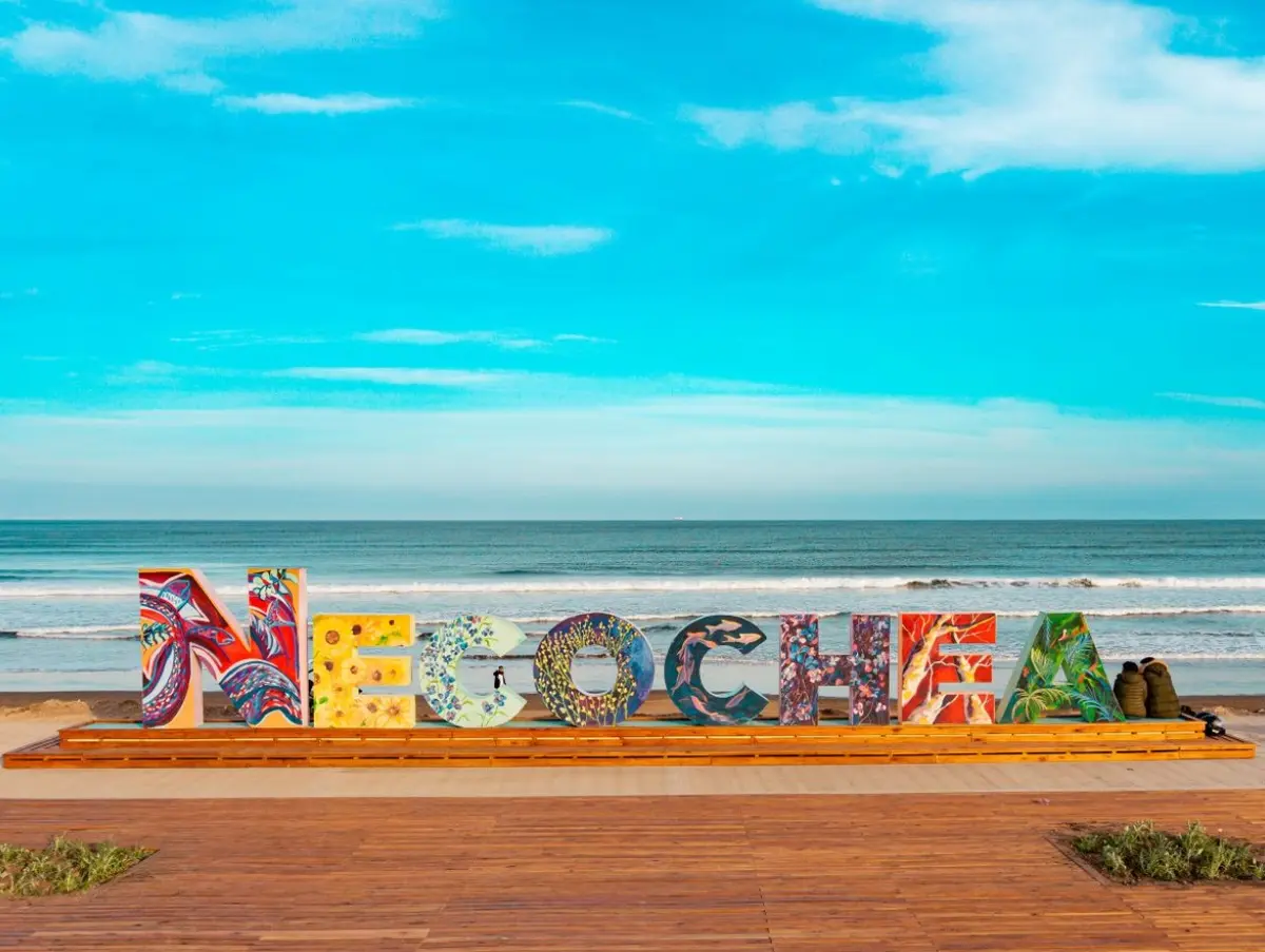 Cartel de letras gigantes con la palabra Necochea pintadas de colores, sobre un deck de madera frente al mar