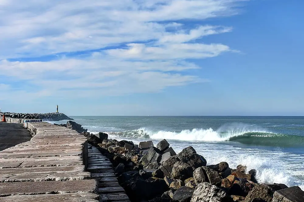 Escollera de bloques de piedra que entra al mar, con una ola rompiendo a la derecha y una pequeña baliza al final
