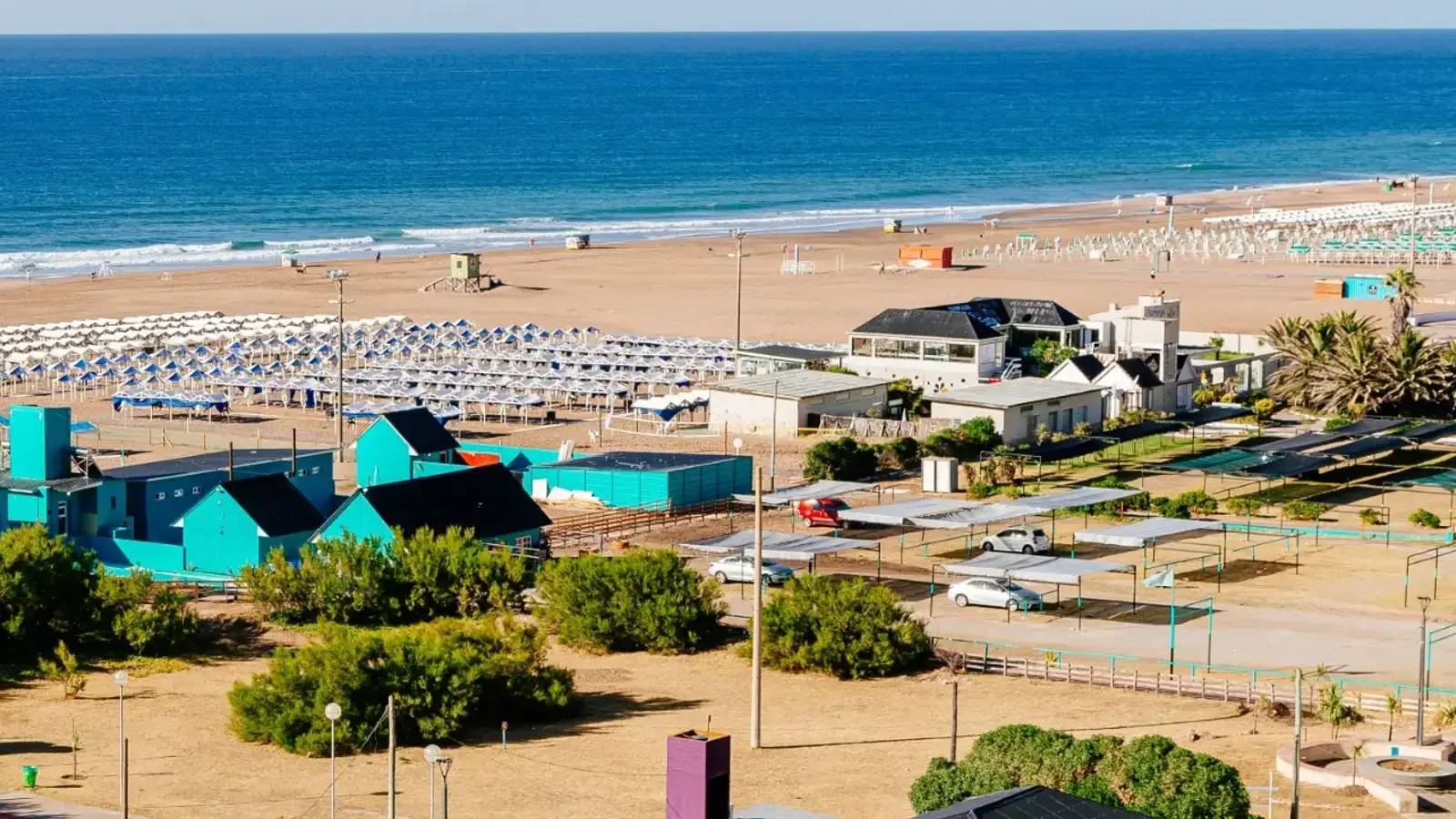 Vista desde lo alto de una playa ancha con hileras de carpas, casillas de guardavidas, construcciones bajas turquesa frente a la arena y el mar azul