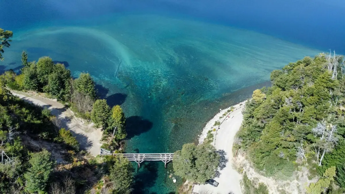 Puente de madera sobre un río de agua transparente que desemboca en un lago turquesa, entre árboles