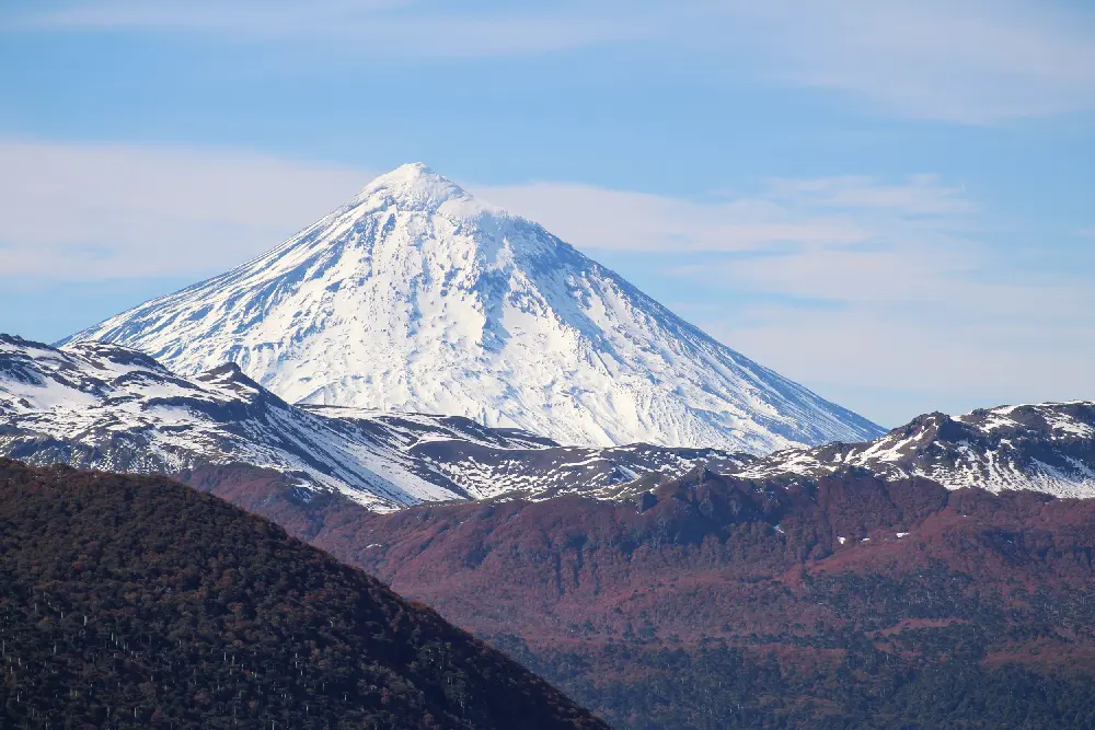 Volcán de cono perfecto cubierto de nieve, detrás de cerros con bosque en tonos rojizos de otoño