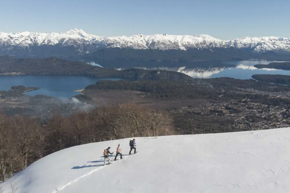 Tres personas caminan con raquetas por una ladera nevada sobre un pueblo, un lago y la cordillera