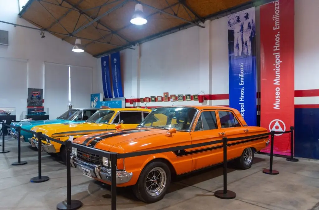 Autos clásicos naranja y amarillo con franjas negras exhibidos en un galpón, con banderas del Museo Municipal Hermanos Emiliozzi