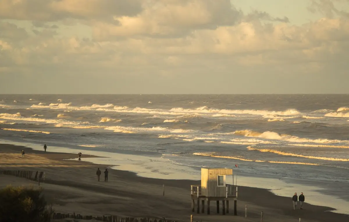 Playa al atardecer con olas espumosas, gente caminando por la orilla y una casilla de guardavidas de madera sobre pilotes