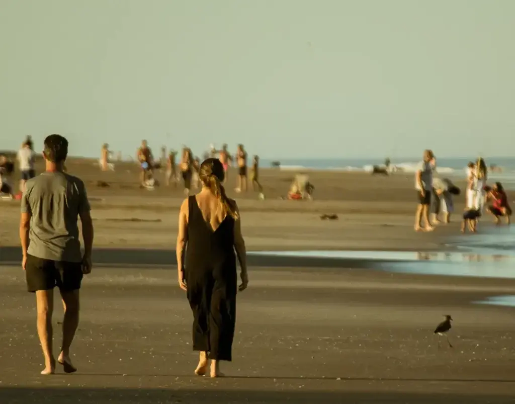 Una pareja camina descalza por la arena húmeda hacia el mar, con otras personas en la orilla y la luz dorada de la tarde