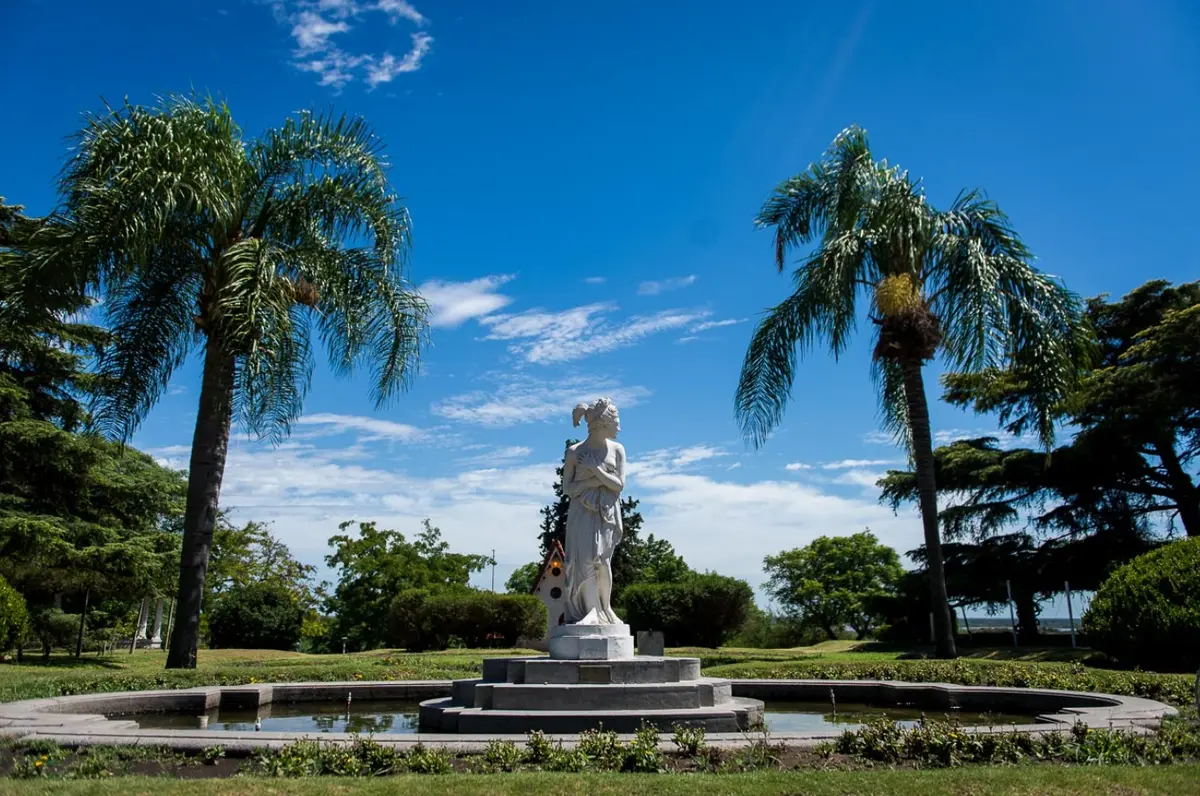 Una estatua blanca sobre una fuente rodeada de palmeras y césped, bajo un cielo azul, con el río apenas visible al fondo