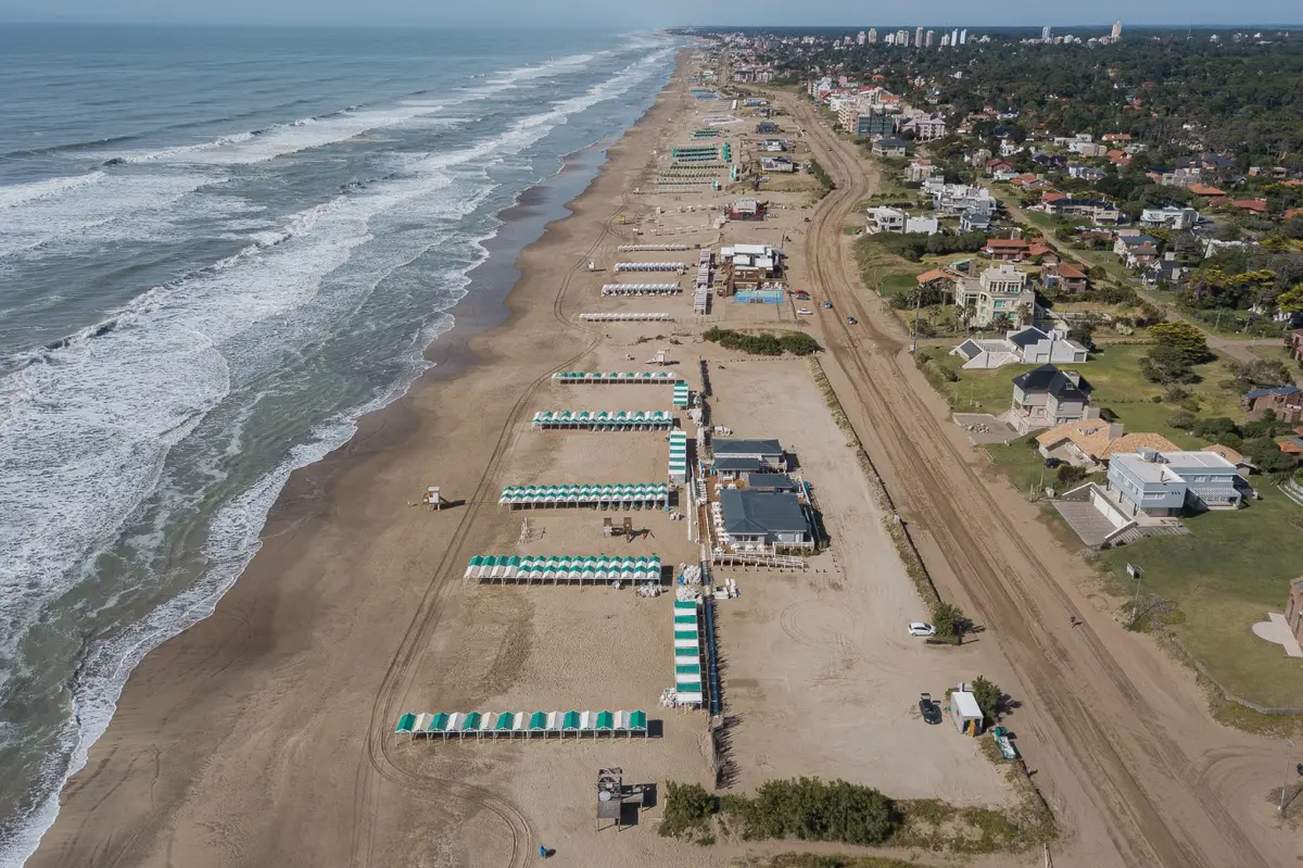 Vista aérea de una playa ancha con hileras de carpas verdes y blancas, balnearios, el bosque con casas a la derecha y edificios altos al fondo