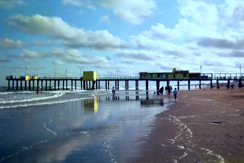 Muelle largo sobre pilotes con construcciones amarillas, gente caminando por la orilla mojada y nubes reflejadas en la arena