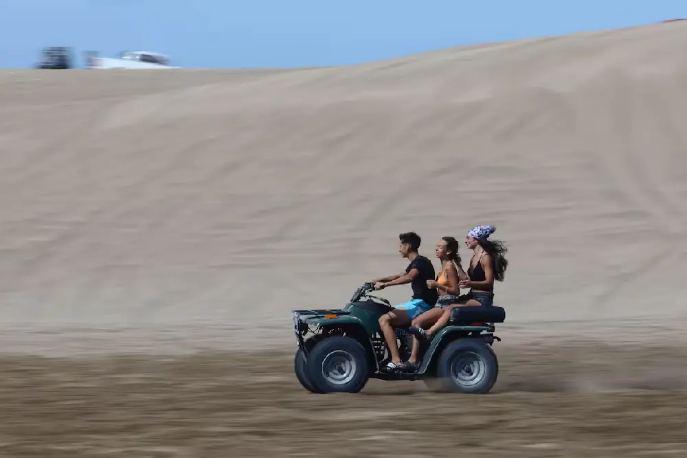 Tres jóvenes andando en un cuatriciclo verde por la arena, con un médano alto de fondo