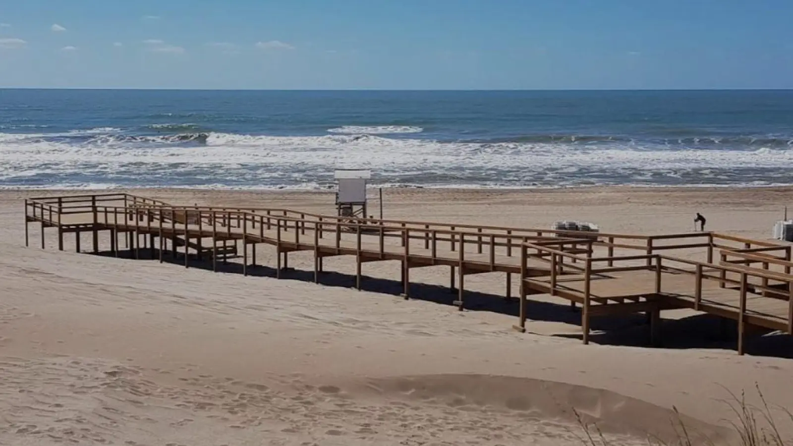 Pasarela de madera que cruza la arena hasta una playa ancha, con una casilla de guardavidas y el mar con olas de fondo