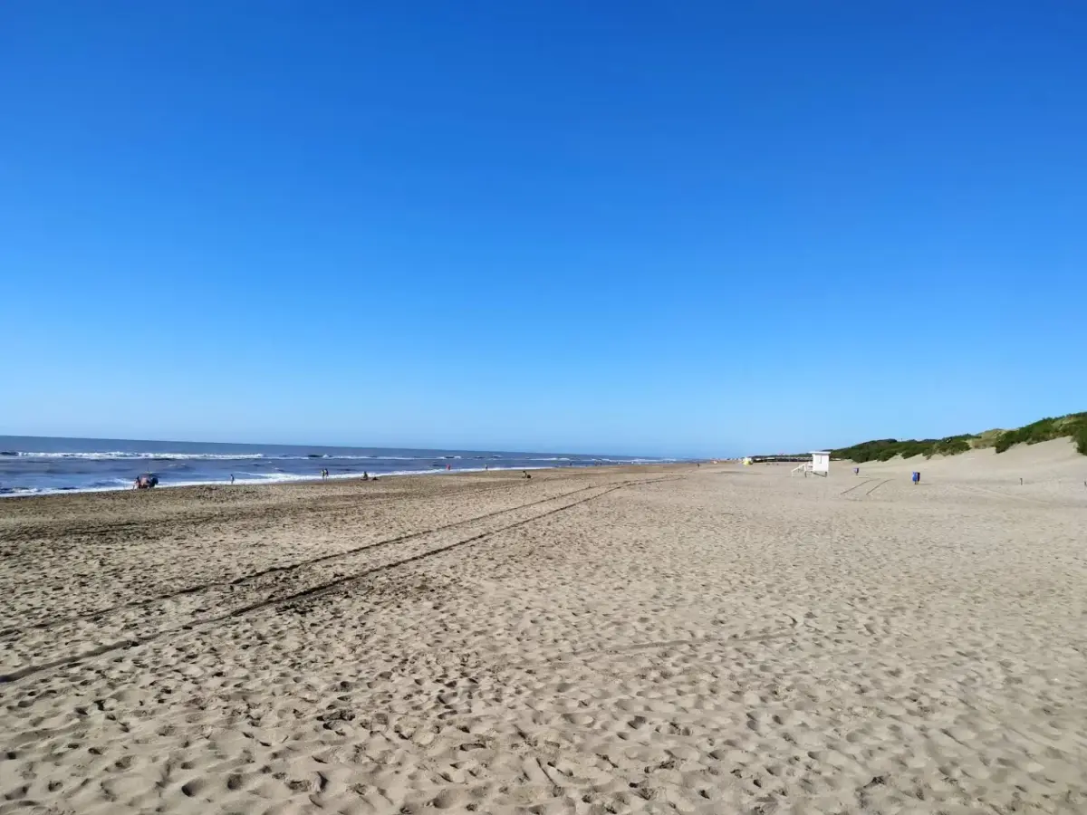 Playa ancha y casi vacía con huellas en la arena, un puesto de guardavidas a lo lejos y médanos con vegetación a la derecha