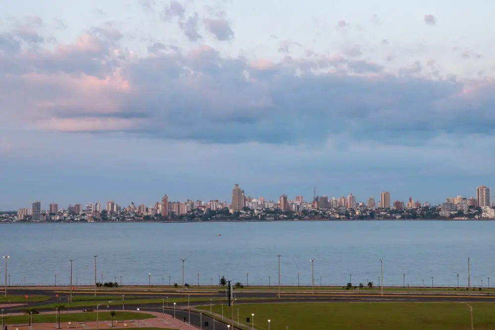 El río Paraná ancho y calmo al atardecer, con una costanera con faroles en primer plano y una ciudad de edificios altos en la orilla de enfrente