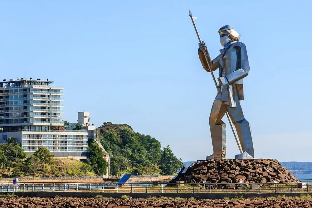 La gran estatua metálica de Andresito con una lanza en la mano, sobre una base de piedras, con un edificio alto y una loma arbolada detrás