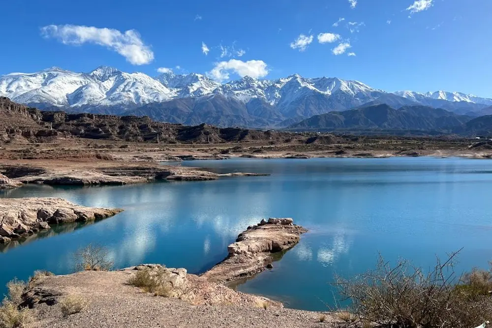 Embalse de agua celeste con orillas de roca clara y arbustos, y la cordillera nevada detrás bajo un cielo azul