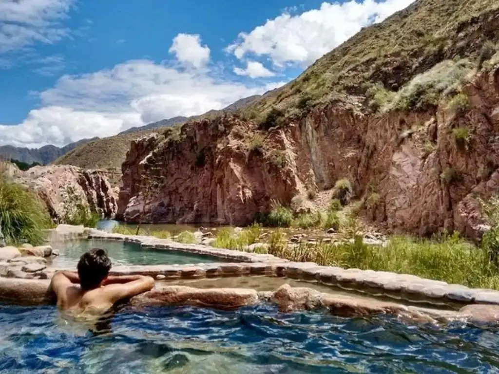 Un hombre de espaldas dentro de una pileta termal de piedra, frente a paredones de roca rojiza y cerros con pasto