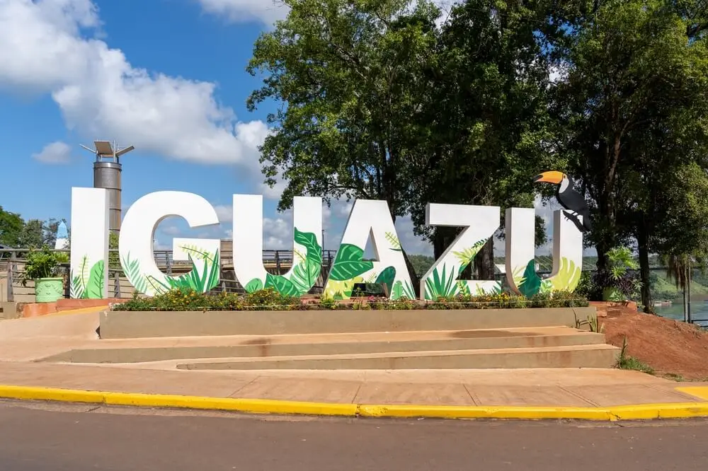 Letras gigantes con el nombre Iguazú decoradas con hojas verdes y un tucán, en una plaza junto al río