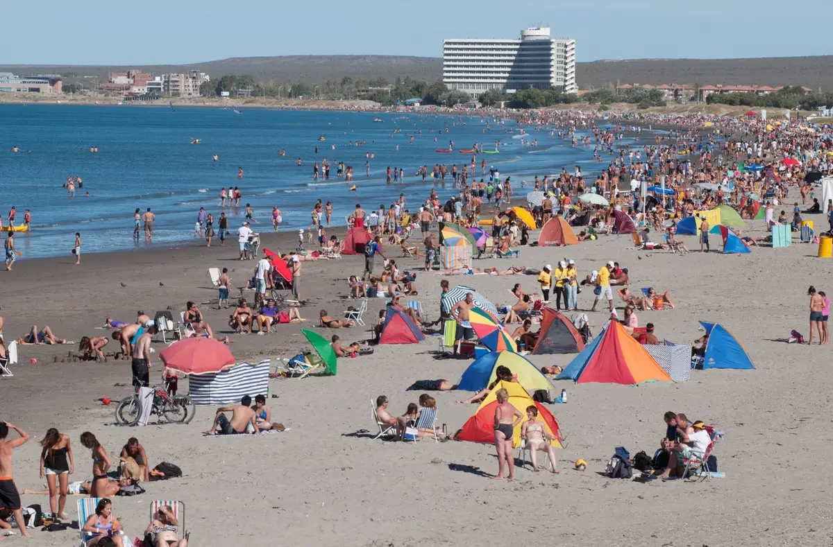 Playa de Puerto Madryn llena de gente con carpas y sombrillas de colores en un día de verano, con el mar calmo a la izquierda