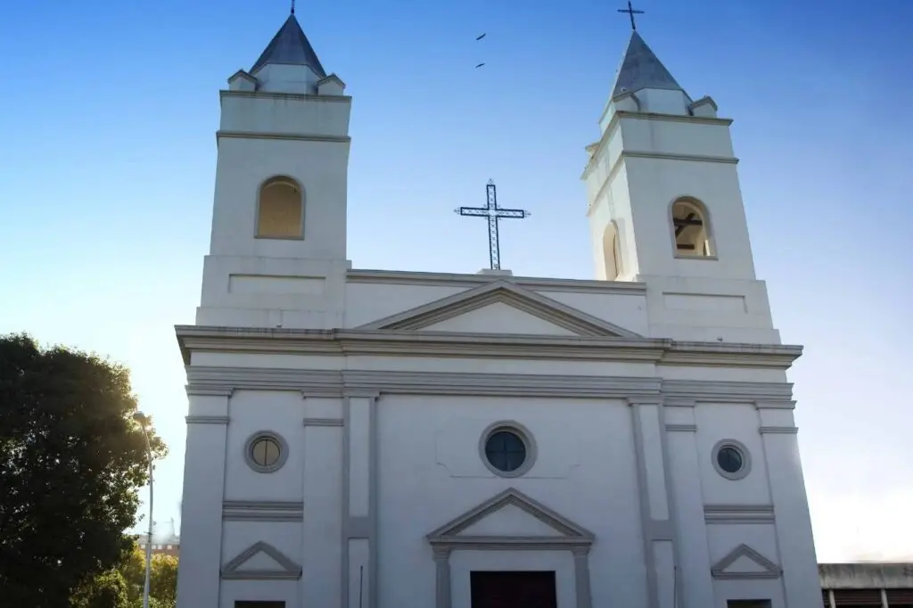 Fachada blanca de una iglesia con dos torres campanario y una cruz de hierro, contra el cielo azul