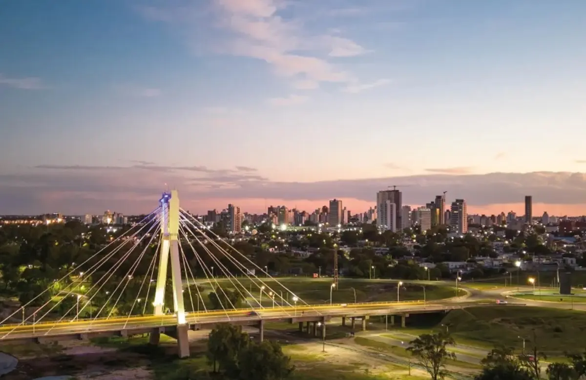 Puente atirantado iluminado sobre una avenida al anochecer, con los edificios de la ciudad recortados contra el cielo rosado
