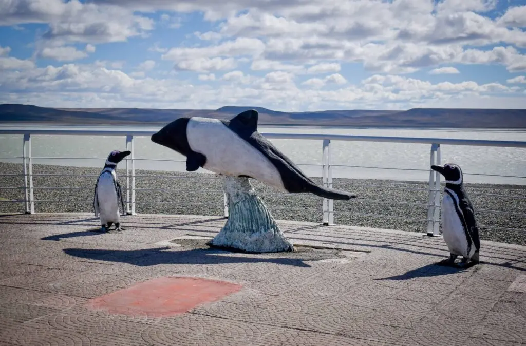 Esculturas de una tonina overa y dos pingüinos de Magallanes en la costanera de Río Gallegos, con la ría y la playa de piedras de fondo