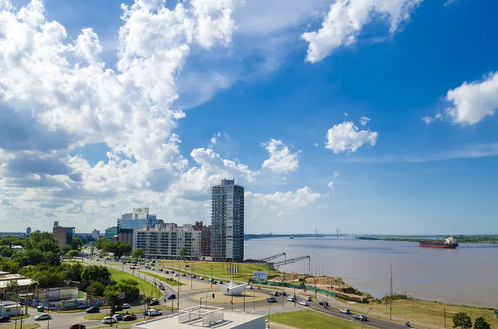 Avenida costanera de Rosario junto al río Paraná, con una escultura blanca con forma de barco de papel en una rotonda, un edificio alto y un buque de carga en el agua