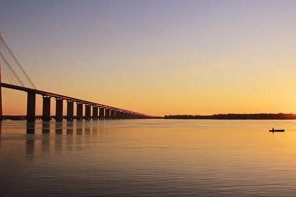 El Puente Rosario-Victoria recortado contra un atardecer naranja sobre el río Paraná, con un pescador en un bote a lo lejos