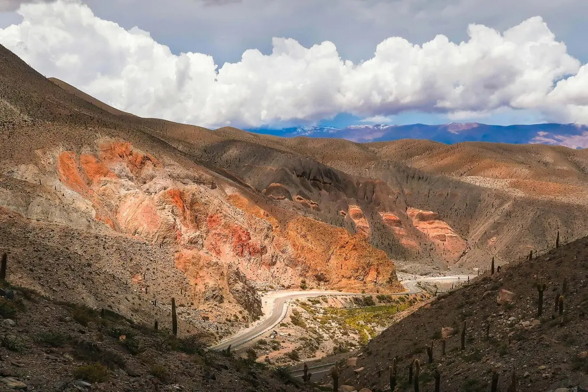 Una ruta serpentea entre cerros ocres y rojizos salpicados de cardones, con picos nevados y nubes blancas al fondo