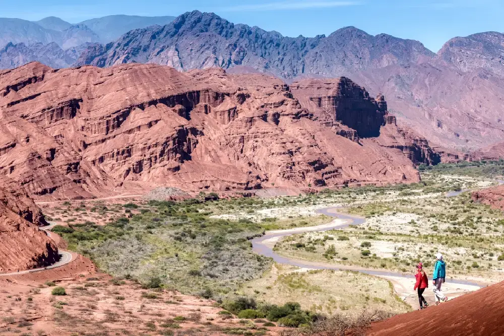 Dos personas caminan por una loma rojiza frente a paredones de roca roja y un río que serpentea por el valle