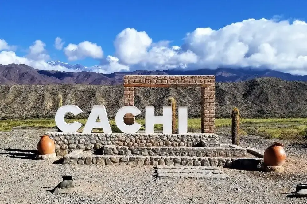 Letras blancas con el nombre de Cachi sobre una base de piedra, con cardones, vasijas de barro y la cordillera nevada detrás