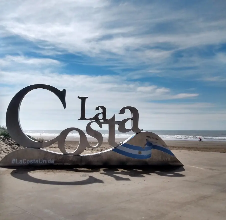Letras gigantes que dicen La Costa sobre una base con la bandera argentina, frente a la playa y el mar bajo un cielo con nubes