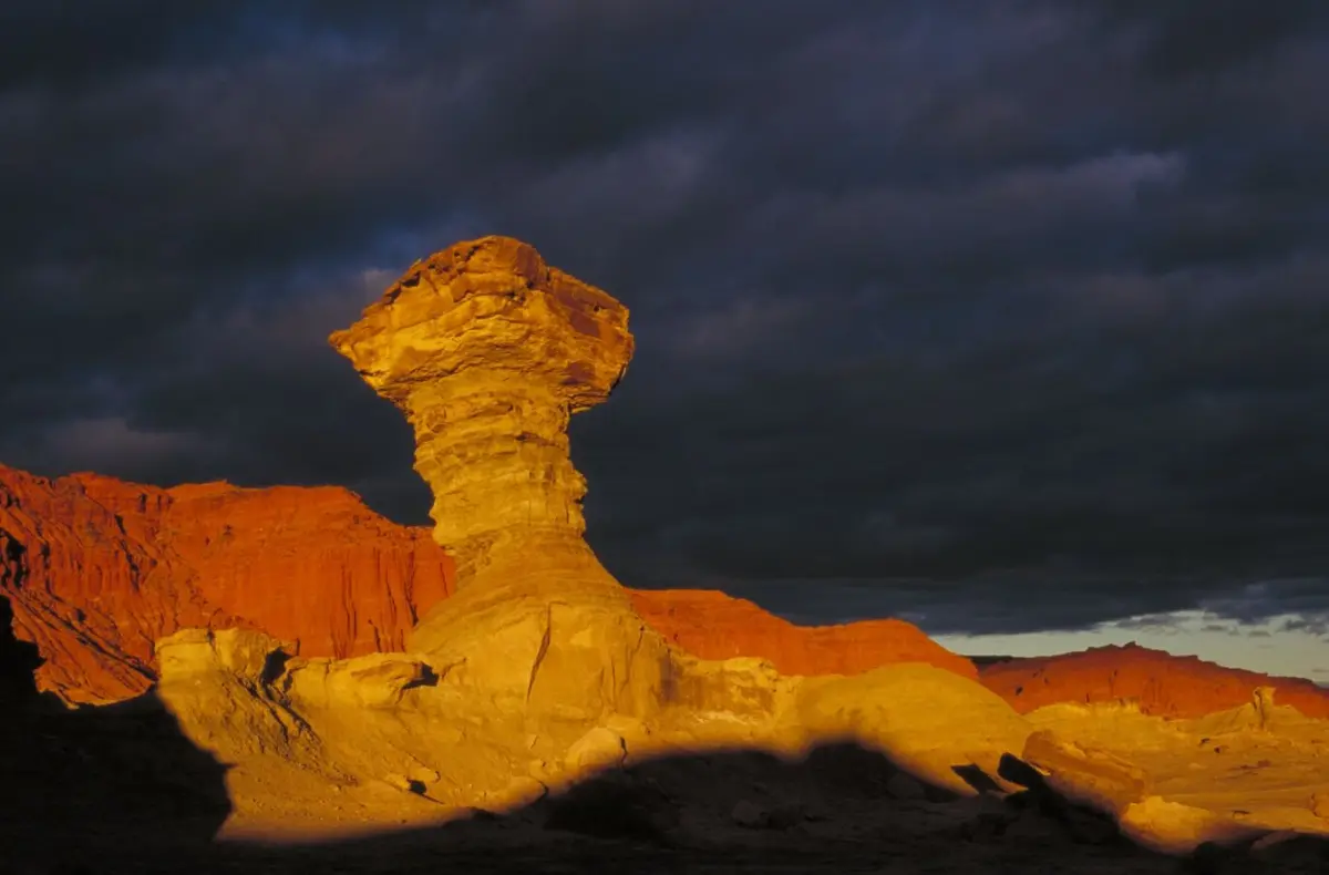La formación de roca conocida como el Hongo, iluminada por el sol, con barrancas rojizas y un cielo de tormenta detrás