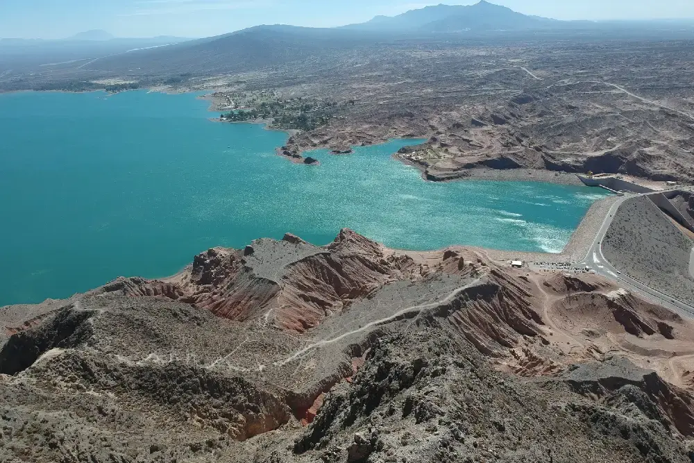 Vista aérea de un dique de agua turquesa entre cerros áridos, con el murallón y la ruta a la derecha