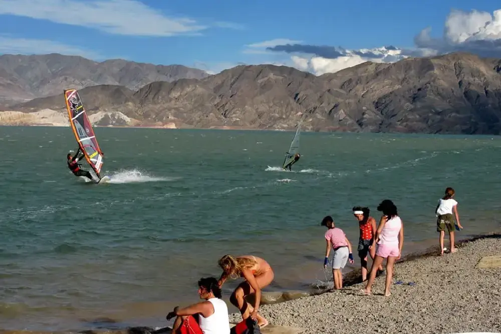 Windsurfistas navegan en un dique rodeado de montañas mientras un grupo de chicas camina por la orilla de piedras