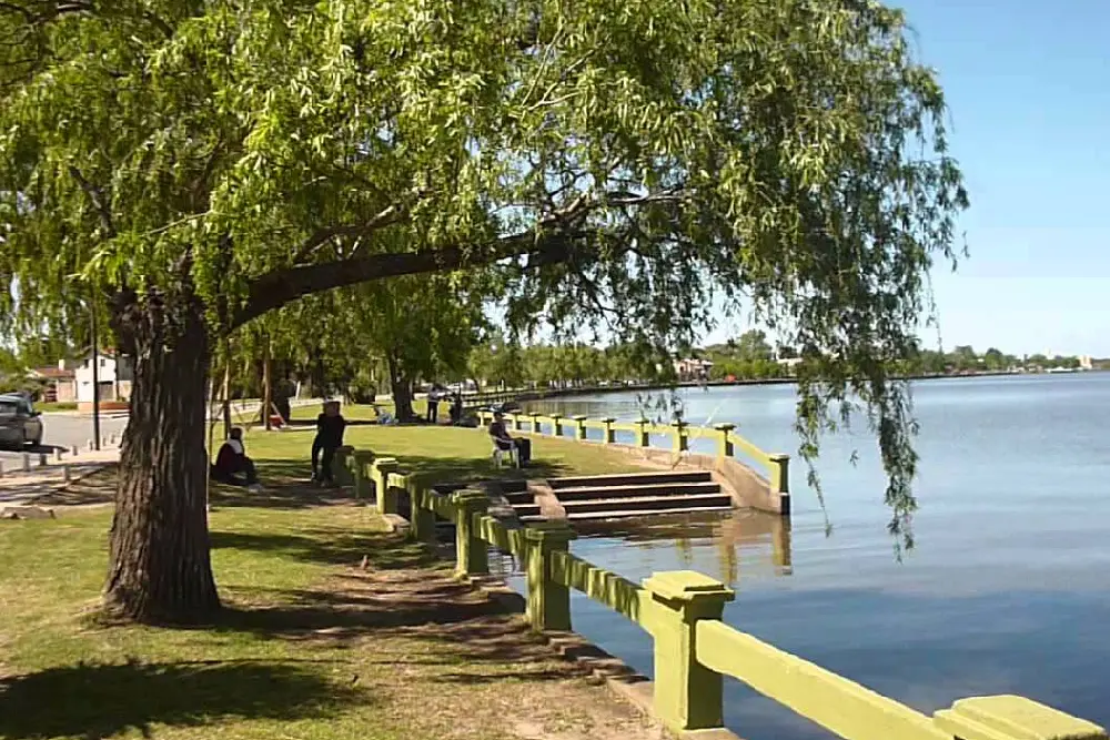 Costanera de la laguna con un sauce sobre el césped, la baranda verde y gente sentada a la orilla del agua