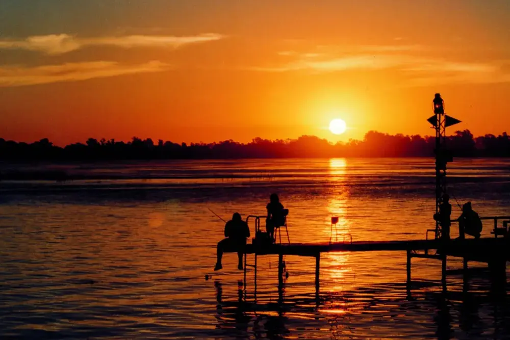 Siluetas de pescadores sobre un muelle de la laguna, recortadas contra el sol naranja del atardecer